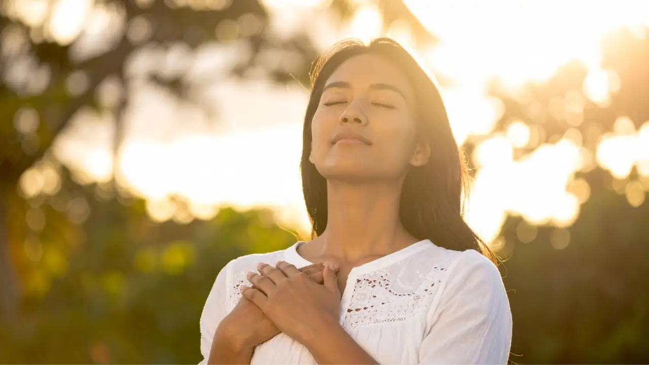 Uma mulher ao por-do-sol, com as mãos unidas sobre o peito com sensação de bem-estar.